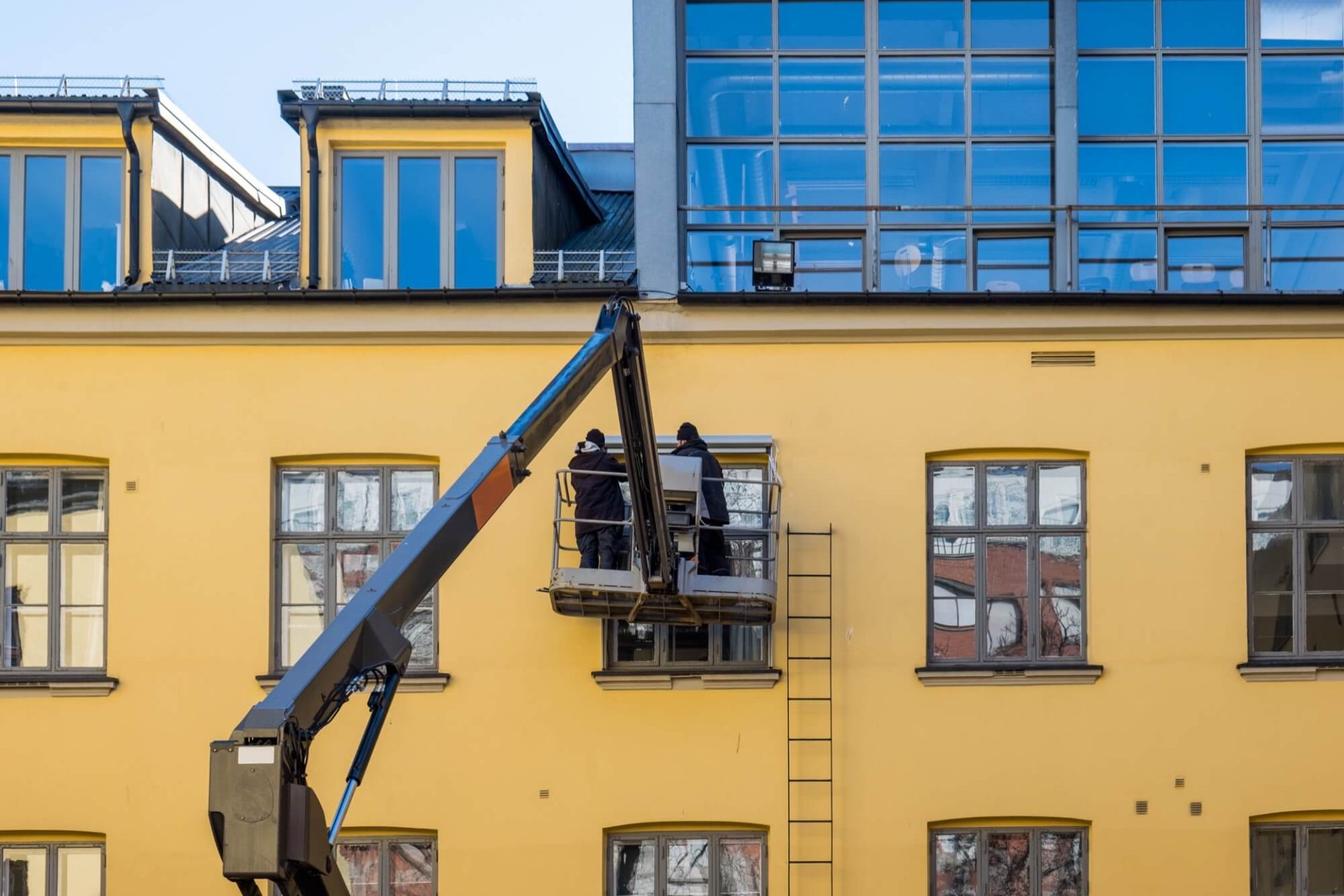 På en markant gul byggnad med generösa fönsterytor ser man arbetare i skylift som renoverar fasaden